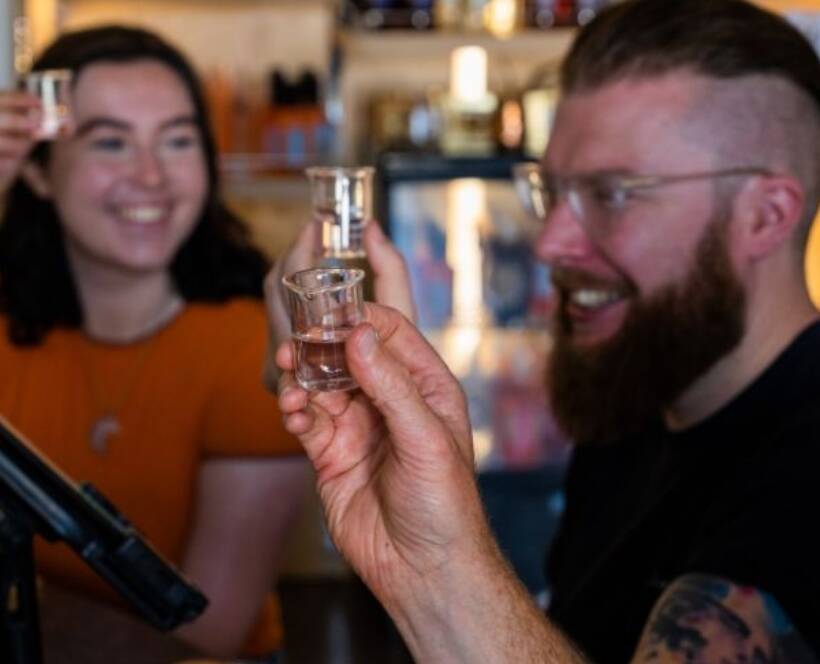 Two people holding shot glasses and smiling at a bar. A hand in the foreground also raises a shot glass. Shelves with bottles are visible in the background.