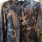 Camouflage-patterned jacket with a central zipper and long sleeves, featuring a mix of brown, green, and gray leaves and branches. It's displayed on a pink hanger against a light-colored background.