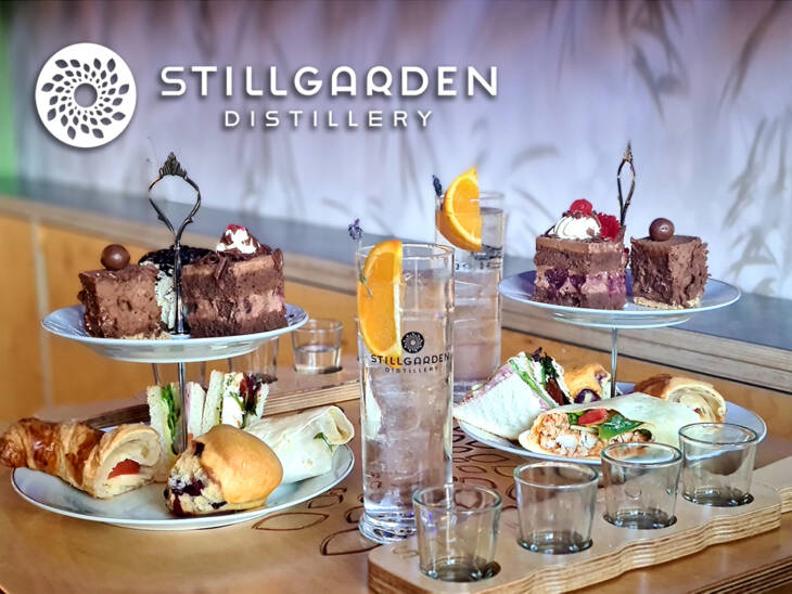 At the Cocktail Masterclass at Stillgarden Distillery, tiered trays of desserts, sandwiches, and pastries accompany a highball glass garnished with an orange slice. A wooden board with empty shot glasses and the distillery's logo create a perfect backdrop.