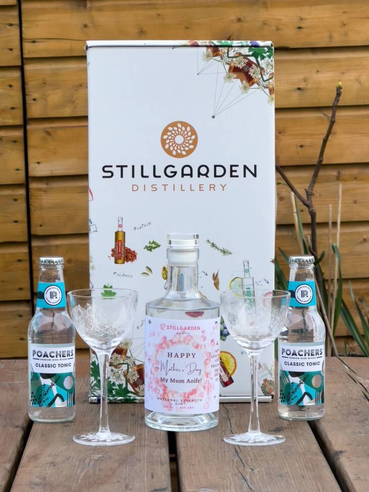 Explore the Personalised Gin Gift Set: a Stillgarden Distillery box with a "Happy Mother's Day My Mum Aoife" pink label bottle, two Poachers Classic Tonic bottles, and two etched glasses, all showcased on a wooden surface.