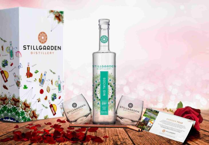 A bottle of Stillgarden Distillery Social Gin is flanked by two branded glasses, with a distillery tour card and rose petals. The allure is heightened by a pink bokeh in the background and a gift box labeled "The Social Box (For Everybody).