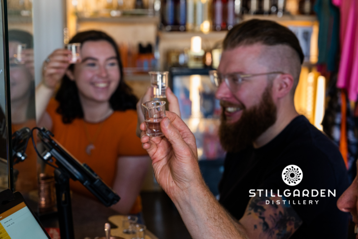 A bearded man and a woman smile while holding up shot glasses in a distillery. Shelves filled with bottles are behind them. A logo reads "Stillgarden Distillery" in the bottom right corner.