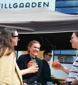 Three people are sitting outdoors under a canopy, holding drinks and smiling at each other. A fourth person is in the background. A building with the word "STILLGARDEN" is partially visible. The setting appears to be a relaxed social gathering.