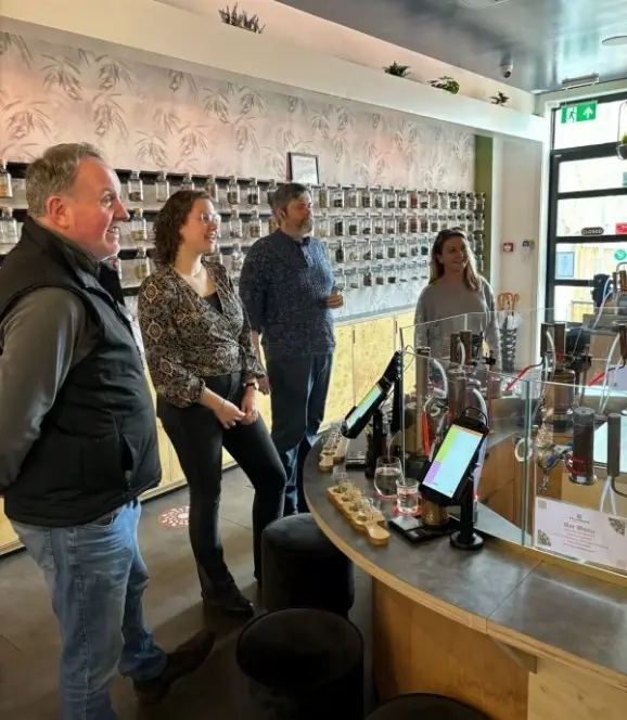 Four people stand inside a modern shop with shelves lined with jars behind them. A counter displays several small items and screens. The atmosphere is casual, with natural lighting coming through a window.