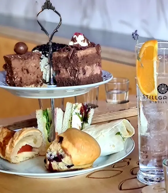 A two-tiered dessert stand holds chocolate cakes, sandwiches, a pastry, and a scone. Next to it, a clear glass with a slice of orange and a sprig of lavender is filled with a sparkling drink. The background shows a wooden table and a blurred wall.