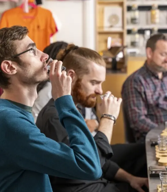 Three people sitting at a table sampling small drinks. The person in the foreground, wearing glasses and a teal sweater, is sipping from a shot glass. Others around the table are also tasting and examining beverages, with various items in the background.