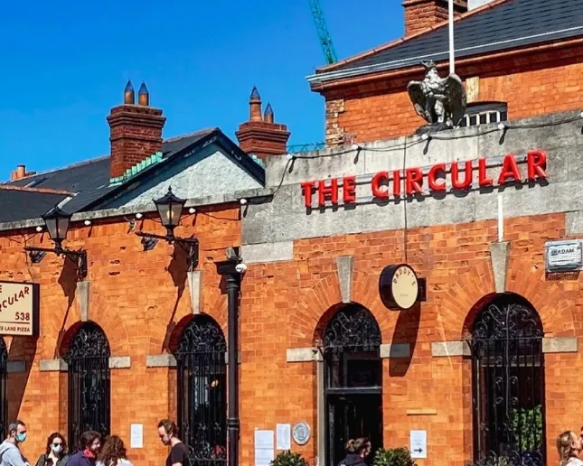 A vibrant scene outside "The Circular" pub, featuring a brick facade and black wrought iron detailing. A small crowd of people walk past, and the sky is clear blue. A bird statue perches above the sign.