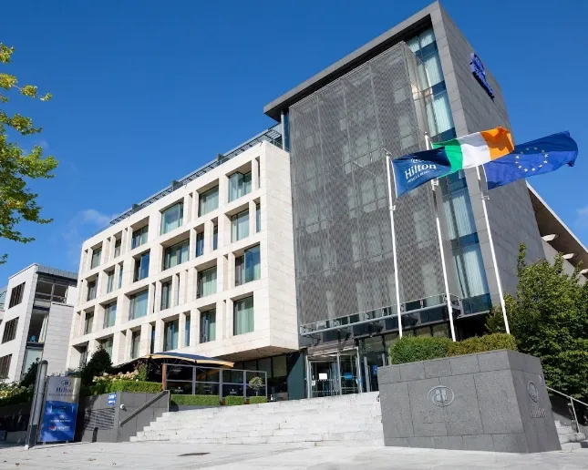 A modern hotel building with beige stone and large glass windows. Flags of the Hilton, Ireland, and the EU are displayed at the entrance. A blue sky is in the background, and the hotel sign is visible on a stone wall near the entrance steps.