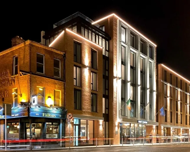 A well-lit street scene at night featuring modern buildings adorned with warm lights. An older brick building with a small bar, "Fallons," is on the left, and the Irish and European Union flags are visible outside a larger structure.