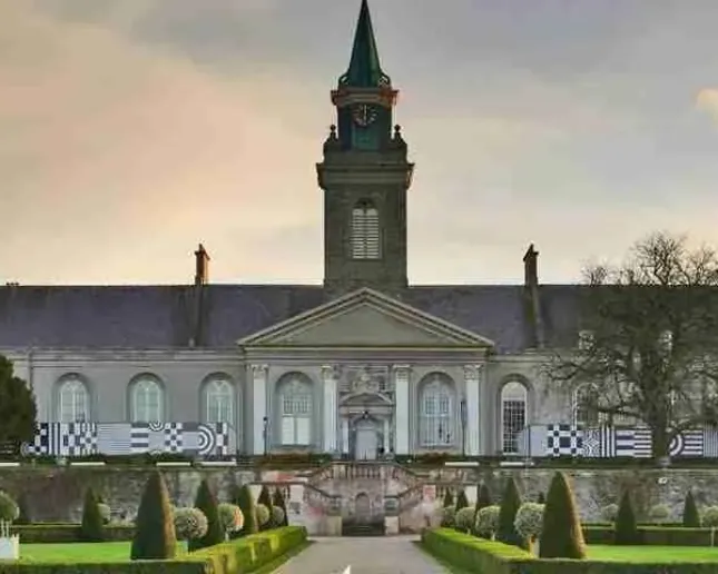 A historic building with a tall clock tower at the center, surrounded by manicured gardens and symmetrical hedges, under a slightly overcast sky. A path leads to the entrance, flanked by decorative flowerbeds and trimmed bushes.