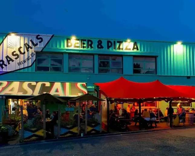 Outdoor view of Rascals Brewing Co. at dusk, with a brightly lit sign for "Beer & Pizza." A group of people are sitting under a red awning at tables in the patio area, enjoying a meal and drinks. The building is teal-colored.