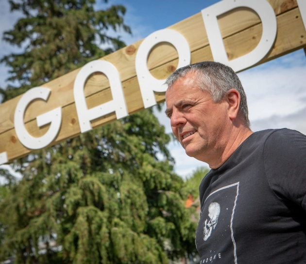 A man wearing a black shirt with a skull design stands outdoors in front of a wooden sign with large white letters. Trees and a cloudy blue sky are in the background.