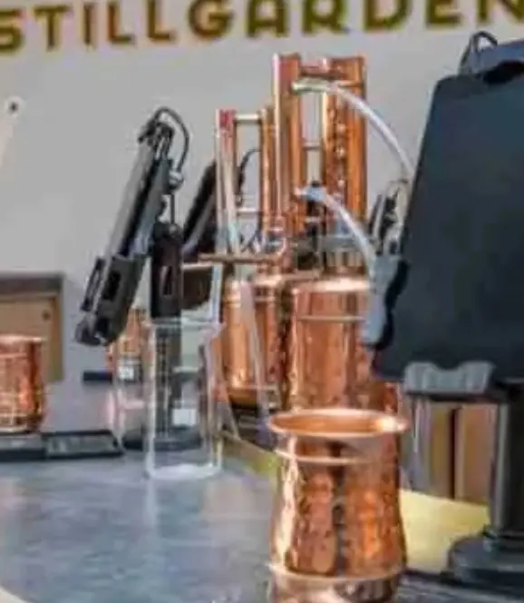 Copper distillation equipment and glass bottles on display at Stillgarden, with a blurred sign in the background.