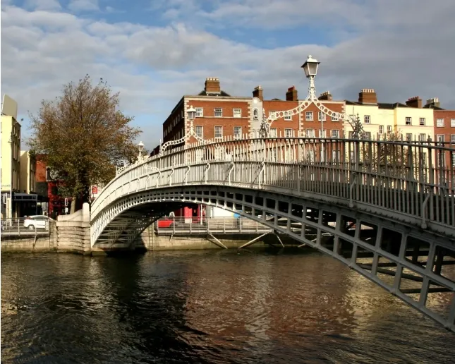 A curved metal pedestrian bridge spans a calm river, with intricate white railings. Behind it, several historic brick buildings with chimneys line the street. A tree stands to the left, and the sky is partly cloudy.