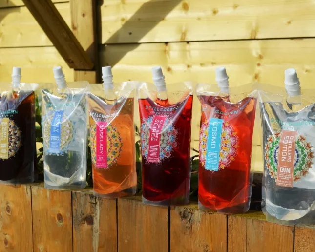 Assorted stand-up pouches containing colorful liquids, likely beverages, are lined up on a wooden surface with a light wooden wall background. Each pouch is labeled differently, indicating various flavors or types.