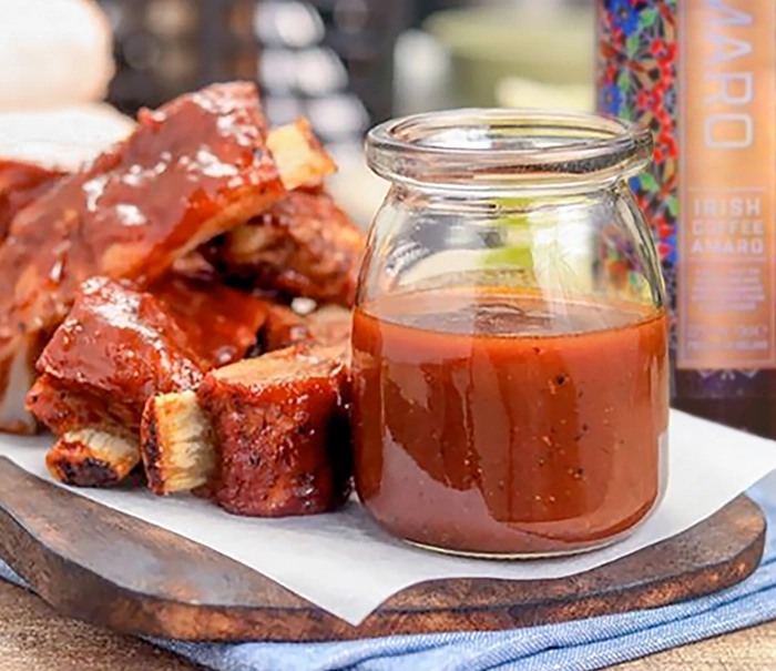 A glass jar of barbecue sauce sits on a wooden board next to a stack of saucy, glazed ribs. A bottle with colorful labeling is visible in the background. The setting is bright and casual, suggesting an outdoor dining experience.