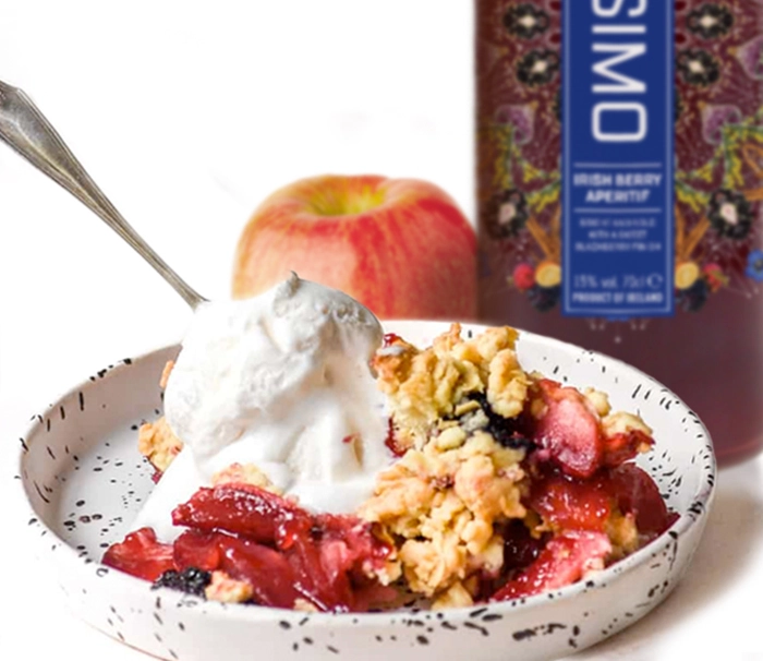 A dessert plate with a fruit crumble topped with a scoop of vanilla ice cream. A whole apple and a bottle of Owisio Irish Berry Aperitif are in the background. The plate is white with black speckles and a fork rests on it.