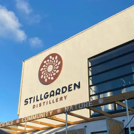 The image shows the exterior of a modern building with the sign "Stillgarden Distillery" featuring a circular floral logo. The words "Science," "Community," and "Nature" are on a wooden beam below. The sky is clear and blue.