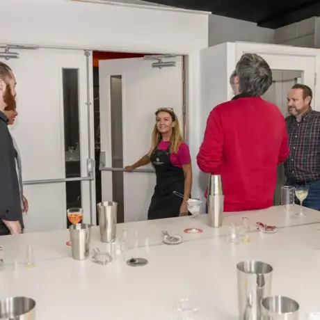 A group of six people gather around a counter with various drink-making utensils. A woman in a pink top stands by the door, smiling. The others, casually dressed, face her, engaged in conversation. Glasses and shakers are scattered on the counter.