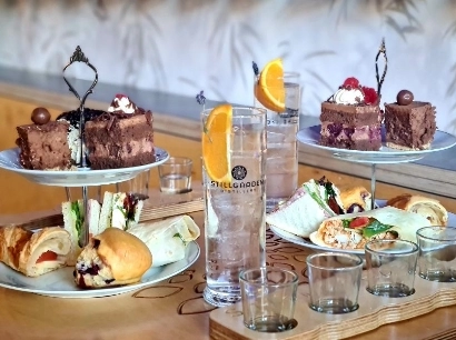 A selection of desserts and sandwiches on tiered trays, including chocolate cakes, wraps, and pastries. Two tall glasses of a clear beverage with orange slices sit alongside, with an empty wooden flight holder in the foreground.