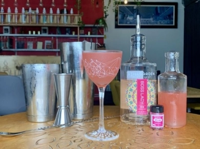 A pink cocktail served in a stemmed glass is on a wooden table. Behind it are cocktail mixing tools, a bottle labeled "Boss Lady," another small bottle, and a shaker. Shelves in the background hold various bottles, with red and dark walls.