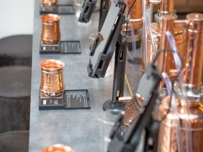 Copper mugs and electronic tablets on display on a gray counter, with various copper items in the background. The scene suggests a modern and industrial aesthetic.