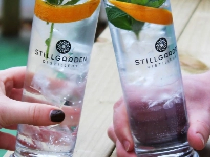 Two hands clink tall glasses filled with ice, clear liquid, mint leaves, and orange slices. The glasses have the Stillgarden Distillery logo. They rest on a wooden surface, suggesting a casual outdoor setting.