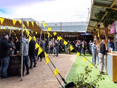 Outdoor gathering at a lively venue with people socializing. Yellow triangular flags are strung along poles. Several food and drink stalls are visible, and people are holding drinks. The setting has a mix of gravel and artificial grass.