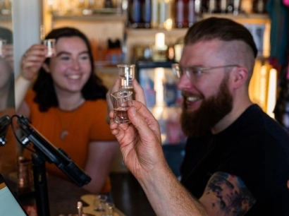 A group of people sitting around a table, holding up small glasses in a toast. One person with a beard is smiling, while another, a woman in an orange shirt, also smiles. Shelves with bottles are visible in the background.