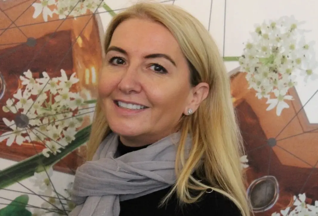 Woman with long blonde hair and a gray scarf, wearing a black top, smiles while standing in front of a wall decorated with abstract geometric and floral designs.