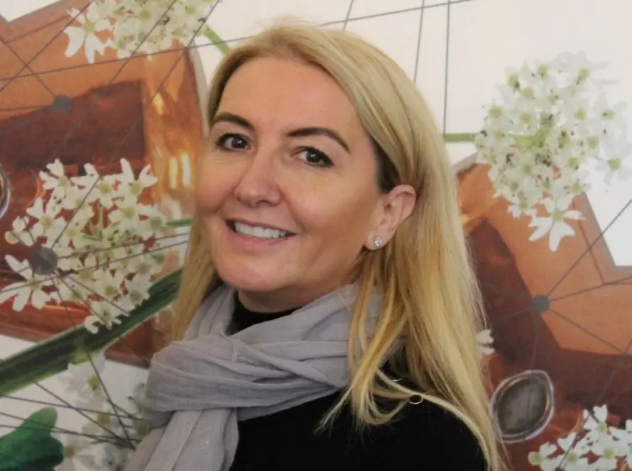 Woman with long blonde hair and a gray scarf, wearing a black top, smiles while standing in front of a wall decorated with abstract geometric and floral designs.