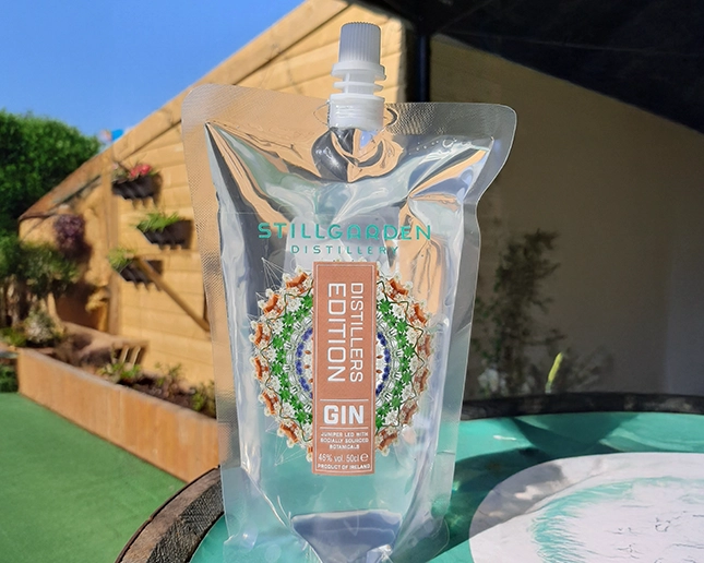 A clear pouch of Stillgarden Distillers Edition Gin stands on a table outdoors, with sunlight shining on it. A garden with plants and wooden fencing is visible in the background.