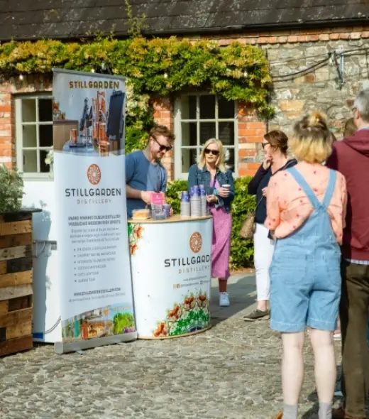 People stand by a booth with "Stillgarden Distillery" banners, sampling drinks outdoors on a sunny day. The setting has a rustic, stone-wall background with climbing plants.