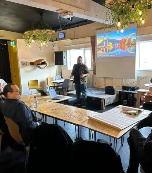 A person stands at the front of a room giving a presentation. There is a colorful slide on a screen behind them. Several people sit at a long, rectangular table, listening intently. The room is decorated with hanging plants.