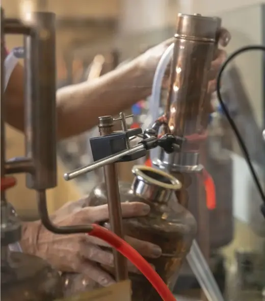 Person operating a copper distillation apparatus with hands adjusting a metal column and connected tubes in a workshop setting. The scene includes glass containers and various equipment.