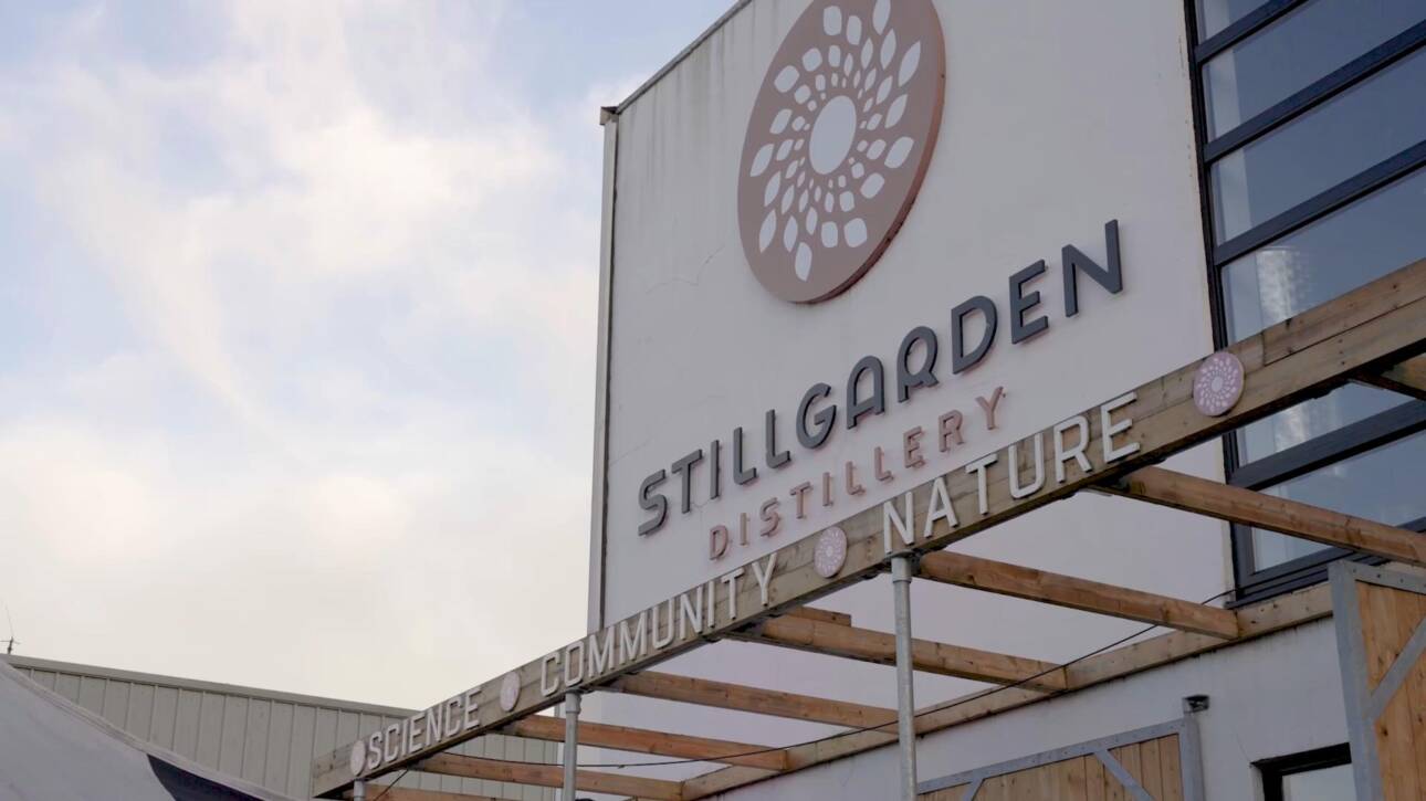 Sign for Stillgarden Distillery mounted on a building, featuring a circular floral logo. Below the logo, the words “SCIENCE,” “COMMUNITY,” and “NATURE” are displayed on beams. Sky with light clouds is visible in the background.