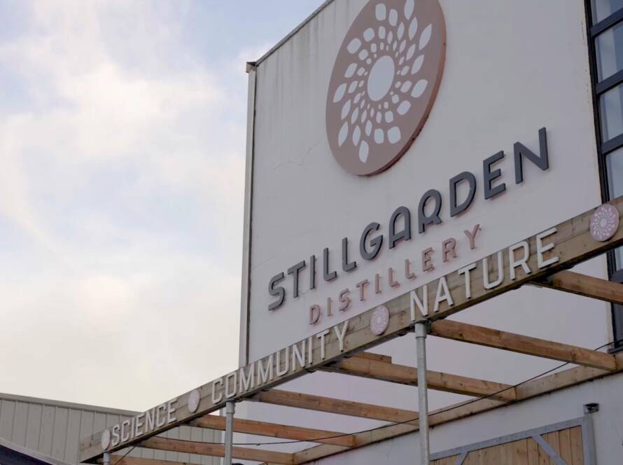 Sign for Stillgarden Distillery mounted on a building, featuring a circular floral logo. Below the logo, the words “SCIENCE,” “COMMUNITY,” and “NATURE” are displayed on beams. Sky with light clouds is visible in the background.